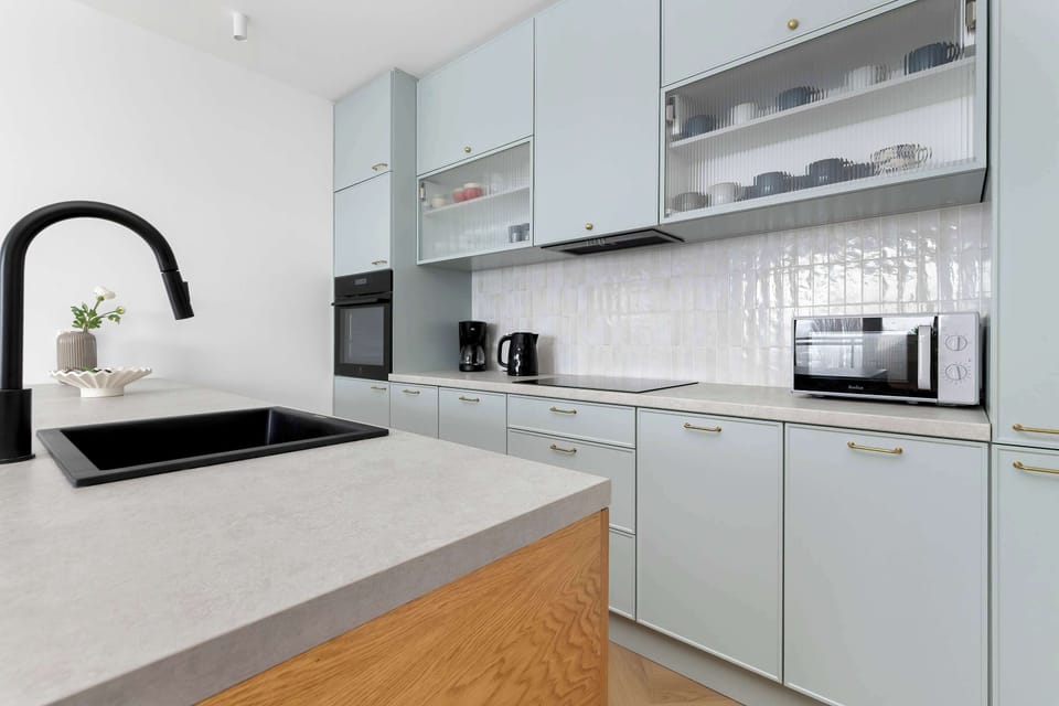 The kitchenette features minimalist blue upper cabinets and a sleek grey countertop. High-end built-in appliances and a black sink highlight the kitchen's functionality and modern aesthetic.