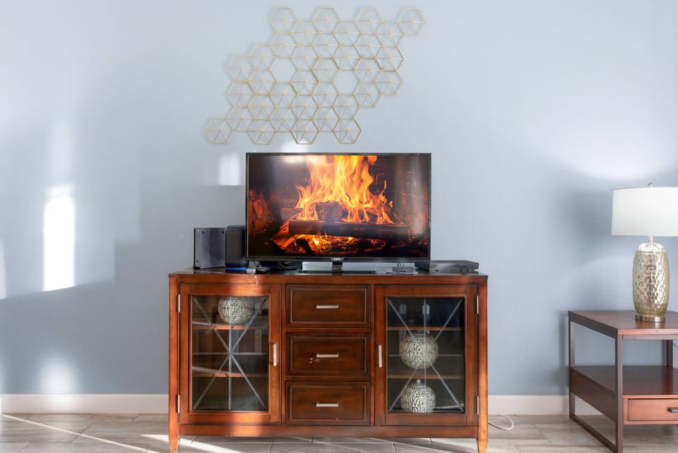 This cozy living area, with its rich wooden media console and soft blue walls, creates a warm ambiance. Sunlight pours in, showcasing decorative elements and inviting you to relax by the flickering fireplace on screen.