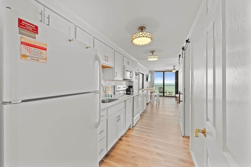 Sleek, modern galley kitchen with bright white cabinetry, updated appliances, and a direct view to the oceanfront balcony — perfect for easy coastal cooking. ️