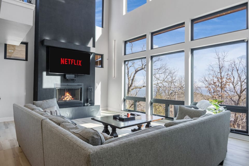 Spacious open concept living room with ample seating, floor to ceiling windows, smart TV, and a wood burning fireplace