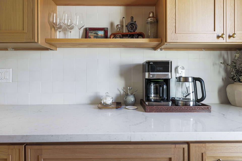 Minimalist coffee nook featuring a sleek brewer and glass kettle on a white marble countertop. Open wood shelving displays glassware against a clean, white tiled backsplash for a modern feel.
