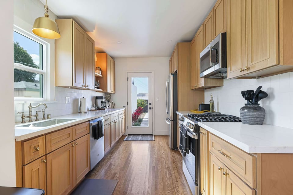 Bright galley kitchen featuring light wood cabinetry, white quartz countertops, and stainless steel appliances. A glass door at the end floods the space with natural light.