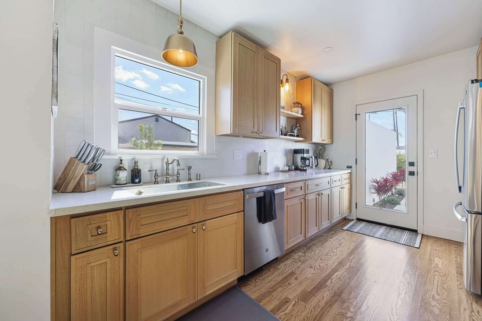 Bright and modern kitchen with wood cabinetry, stainless steel appliances, and a sleek dishwasher. Large windows and a glass door flood the space with natural light for an airy cooking experience.