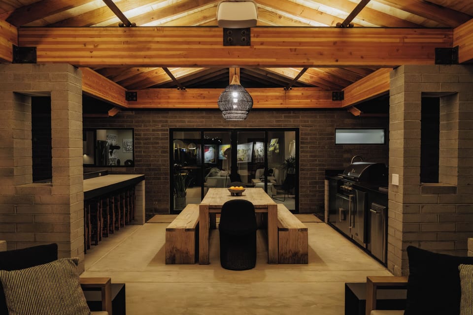 A spacious covered outdoor pavilion with exposed wood beams, ambient lighting, a built-in grill station, bar seating, and a substantial rustic dining table—creating a warm, open-air setting that feels both refined and inviting.
