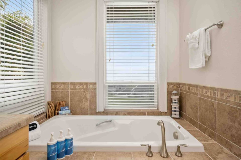 A deep built-in soaking tub positioned beneath two large windows with horizontal blinds, surrounded by warm neutral tile trim.