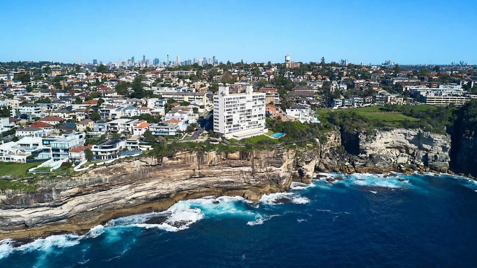 Iconic Cliffside Coastal Enclave

A spectacular aerial panoramic capturing a prominent white architectural residential building perched directly on a rugged sandstone cliff face, looking out over crashing deep-blue ocean waves with a distant city sky