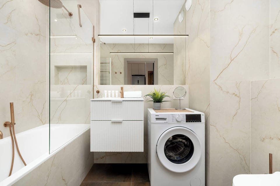 The contemporary bathroom is finished in marble-effect tiles and includes a bathtub with a glass screen. A vanity unit and washing machine maximize the space's utility.