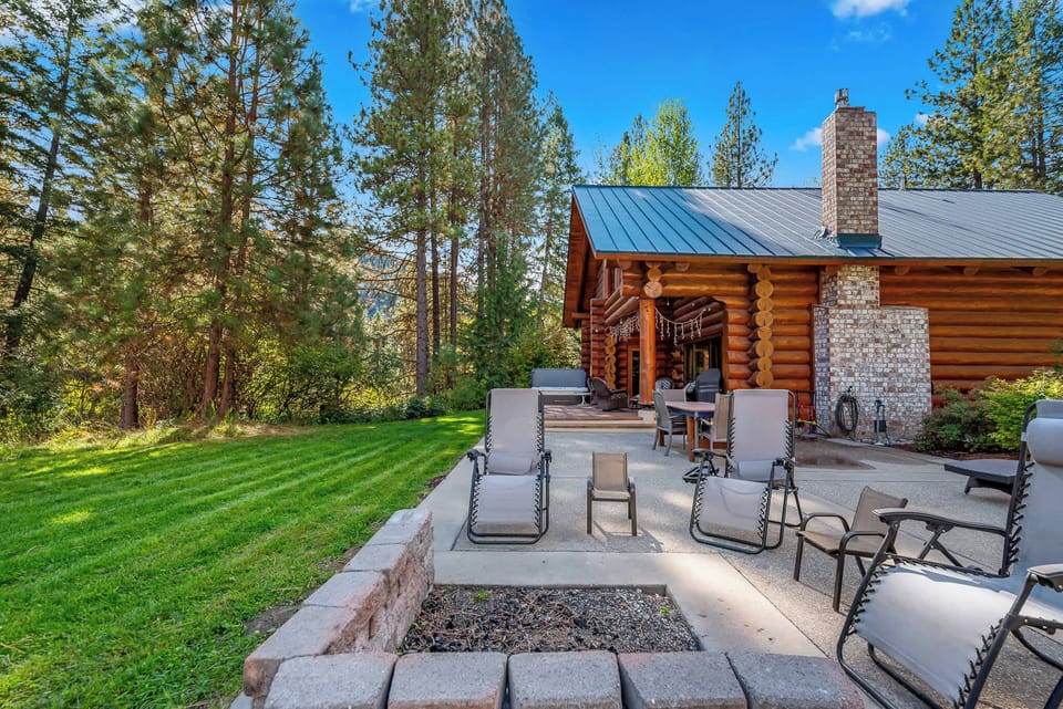 A charming log cabin retreat nestled among towering pines, featuring a spacious outdoor patio with lounge chairs and a stone fire pit—perfect for soaking up the sun or gathering under the stars.