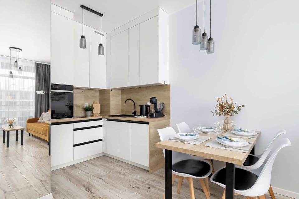 A compact kitchenette and dining corner with white cabinetry and wooden accents. A small table with chairs is placed next to the kitchenette counter. Pendant lights hang from the ceiling.