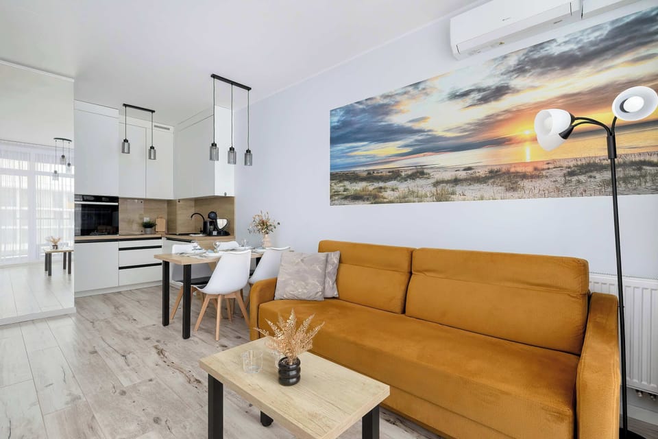 An open-plan living space combining the lounge and kitchen area. A mustard sofa sits near a wooden coffee table, and white kitchen cabinets line the wall. A large landscape painting decorates the room.