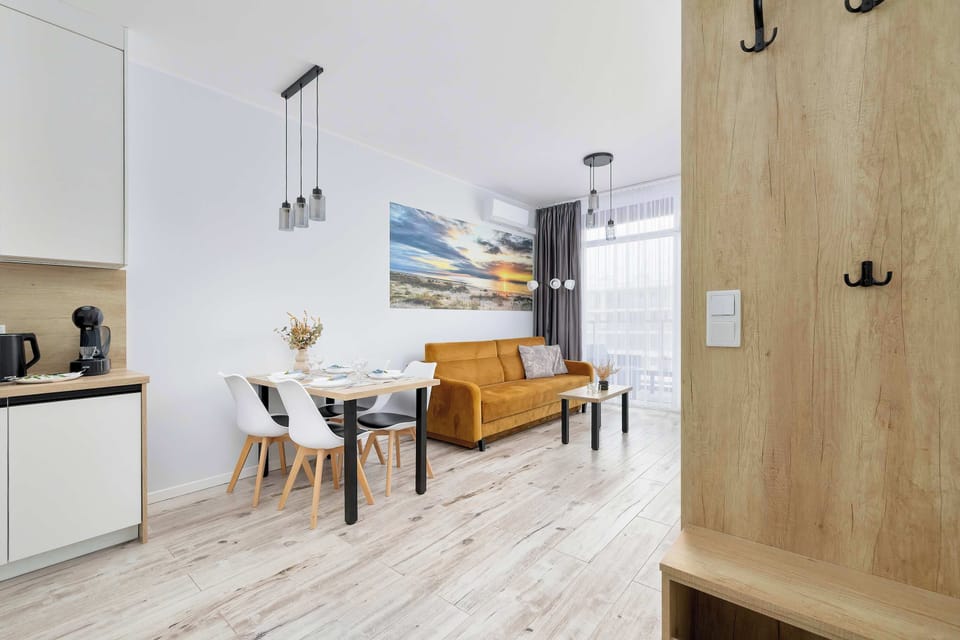 A hallway perspective leading into the open living and dining space. Wooden doors and light flooring create continuity. The mustard sofa is visible in the distance.