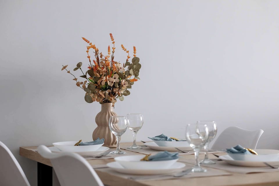 A neatly arranged dining table with elegant glassware and a vase of dried flowers as a centerpiece. Light-colored walls and simple décor create a calm atmosphere. The styling emphasizes attention to detail.