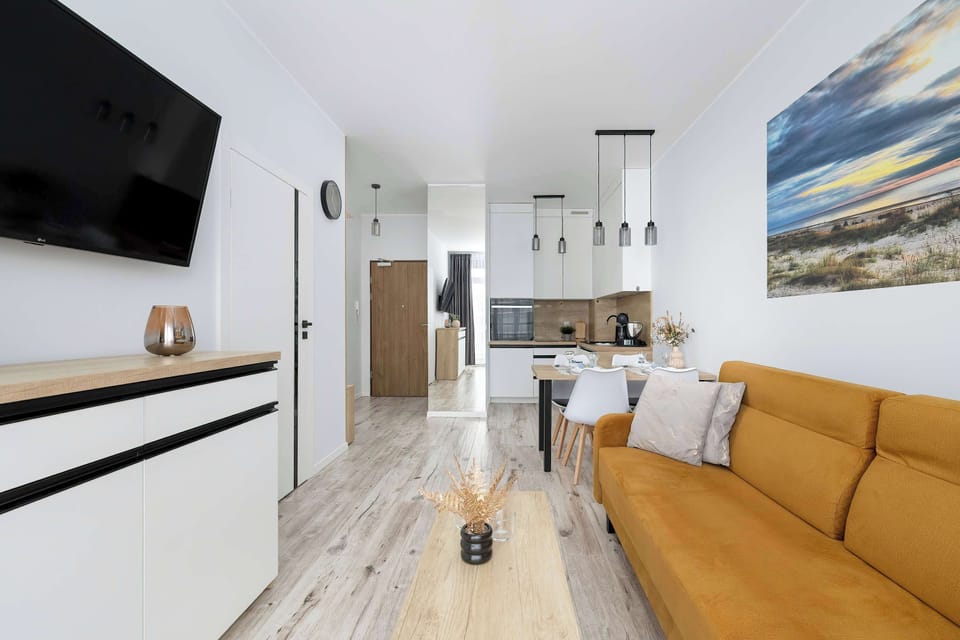 A view from the living room toward the kitchen area with a mounted TV on the wall. The mustard sofa and wooden flooring create a warm ambiance. The open layout enhances functionality.