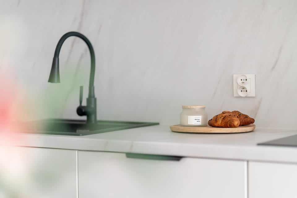 The kitchen sink area features a modern black faucet and a clean white countertop against a marbled wall. A small wooden board with a snack sits nearby, emphasizing a welcoming, lived-in feel.