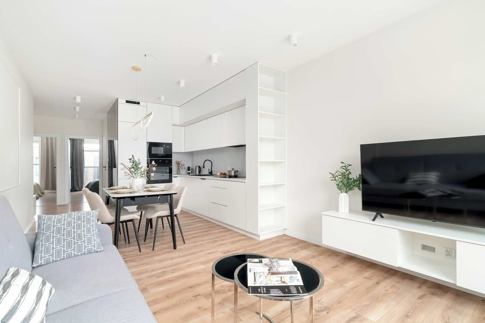 The overall view of the bright living room shows the seamless transition between the lounge area and the dining space. A flat-screen TV is mounted on a white unit, facing the comfortable seating area.