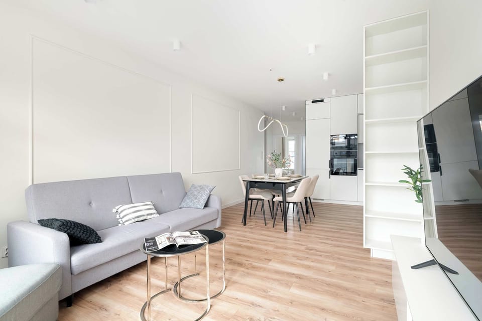 An open-concept living area displays a grey sofa and a modern coffee table with a glass top. In the background, a dining table and a glimpse of the kitchen area are visible, showing a functional layout.