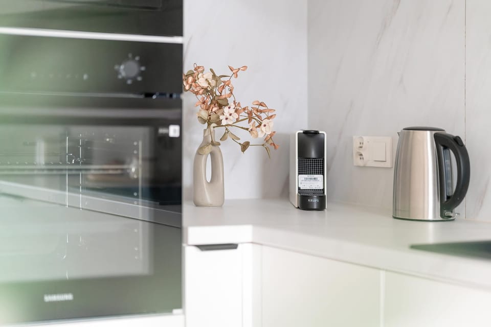 A kitchen counter holds modern appliances including a black built-in oven and a stainless steel kettle. A small vase with dried flowers adds a decorative touch to the functional workspace.