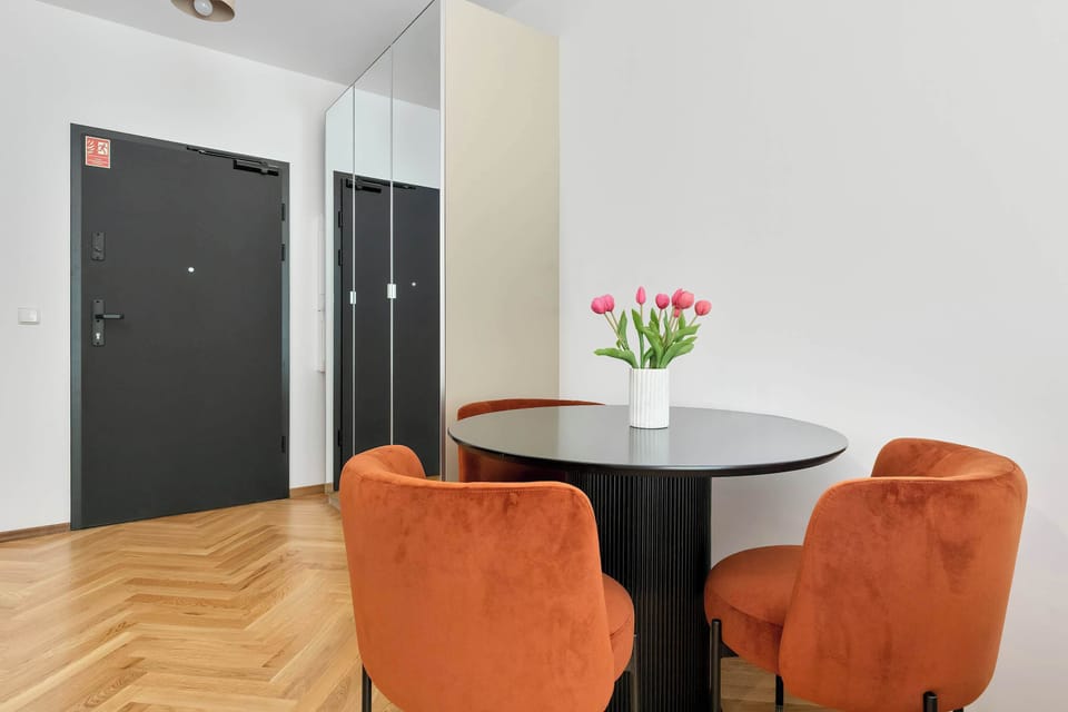 A small dining setup with a black round table, two orange chairs, and a vase of pink flowers, situated near a dark entrance door. The wooden flooring adds warmth to the space.