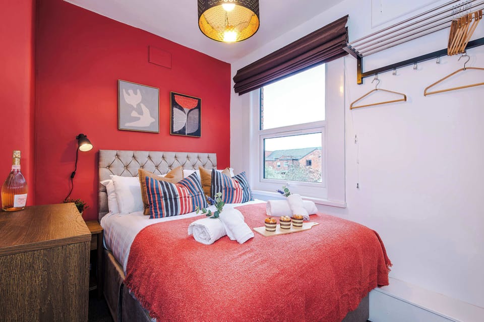 Compact bedroom with a double bed featuring a tufted headboard, red accent wall, and wooden dresser. Includes bedside lamp, framed artwork, and window with Roman shade, bright, cozy space designed for comfort and style.
