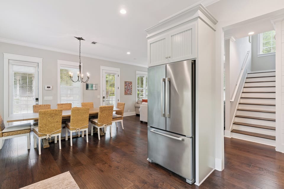Your open-concept kitchen flows seamlessly into a sunny dining area with woven chairs that bring coastal charm and warm wood floors throughout your entire space.