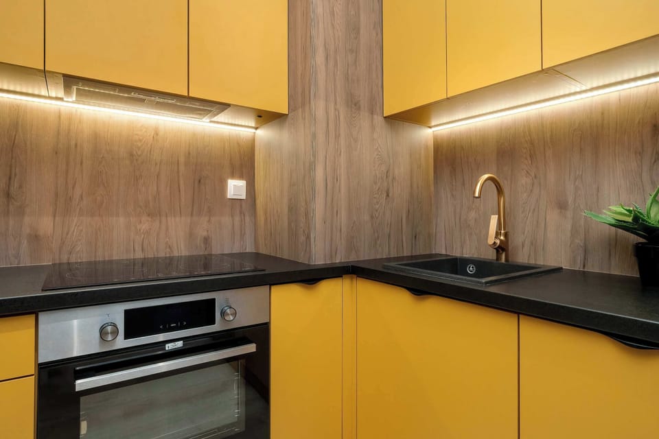 A close-up of the kitchen countertop with a built-in oven and stovetop. The combination of yellow cabinets and wood-textured backsplash creates a warm ambiance. Small potted plants decorate the counter.