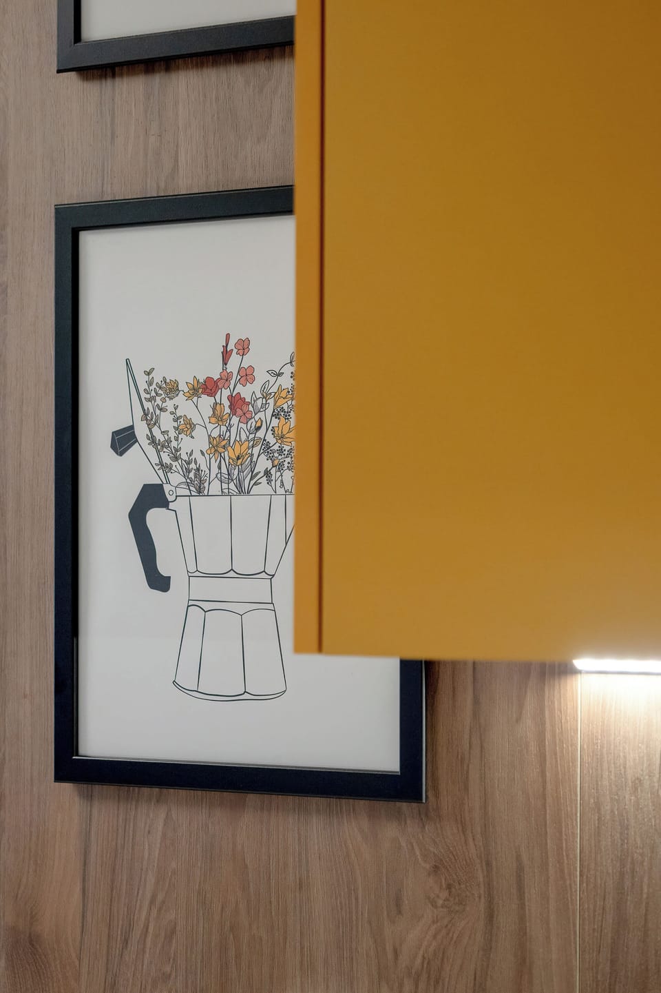 A decorative detail of framed artwork mounted on a wooden wall panel. A yellow cabinet edge is visible nearby. The artwork adds personality to the interior.