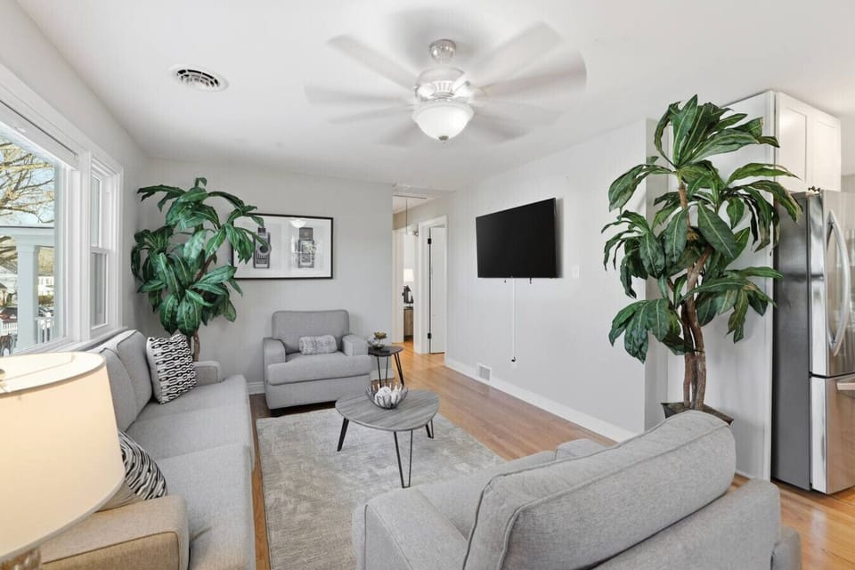 Bright, inviting living room with a large flat-screen TV, comfortable sofa, and two accent chairs—perfect for relaxing after a day out. The oversized window brings in natural light, and the open layout flows into the dining area for easy gathering.
