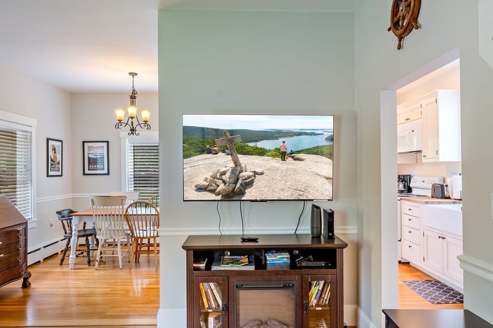 Modern entertainment in a 1900s cottage. Our new 55-inch Roku TV sits above an electric fireplace, flanked by a dining area and kitchen. Spot the Acadia Mountain summit on screen—a peak you can hike just minutes from this Hall Quarry home.
