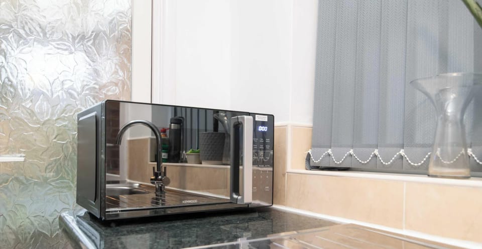 A sleek, mirrored microwave sits on a dark countertop, reflecting the surrounding kitchen and a window with grey blinds.