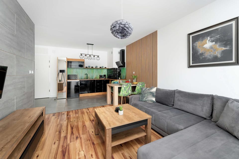 A spacious living room area features a deep teal L-shaped sofa and a minimalist black coffee table. Exposed wooden beams and a bright skylight create an airy, loft-like atmosphere over the light wood flooring.
