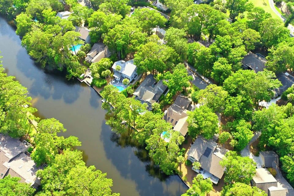 Leafy canopies shade this peaceful waterfront neighborhood, where shingle roofs and private docks meet the calm, dark water. Morning brightness plays across the lush gardens and turquoise pools, creating a quiet sanctuary for your next getaway.