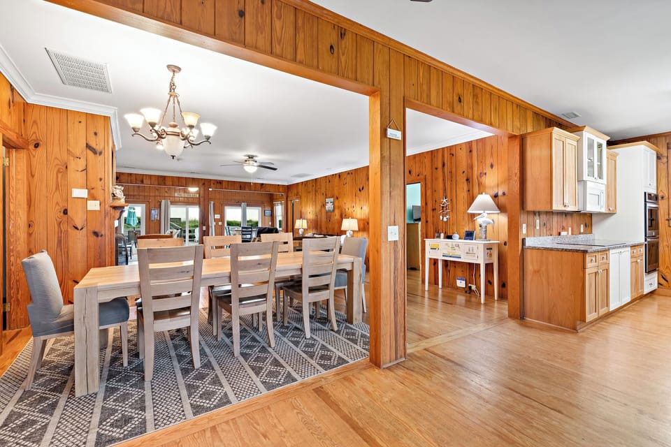 Warm Oak Island NC dining room with wood-paneled walls, large wooden table for six, chandelier lighting, patterned rug, and open access to the kitchen and deck through glass doors.
