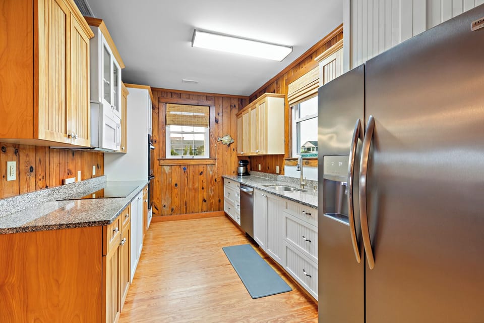 Charming Oak Island NC kitchen with stainless steel refrigerator, granite countertops, white cabinets, wood-paneled walls, double sink under window, dishwasher, microwave, and bright overhead lighting.