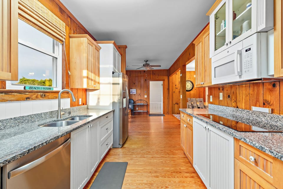 Charming Oak Island NC kitchen with stainless steel refrigerator, granite countertops, white cabinets, wood-paneled walls, double sink under window, dishwasher, microwave, and bright overhead lighting.