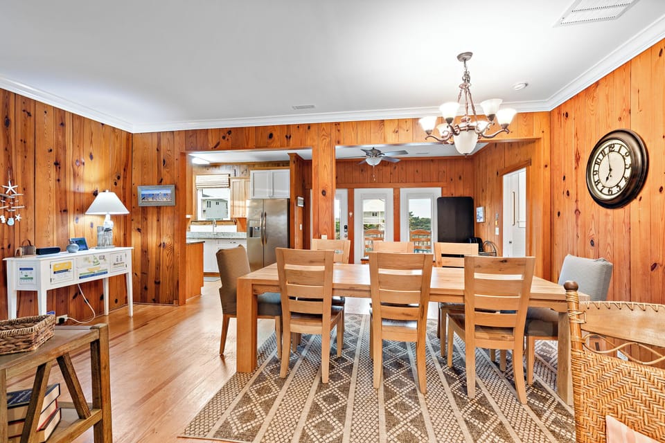 Warm Oak Island NC dining room with wood-paneled walls, large wooden table for six, chandelier lighting, patterned rug, and open access to the kitchen and deck through glass doors.