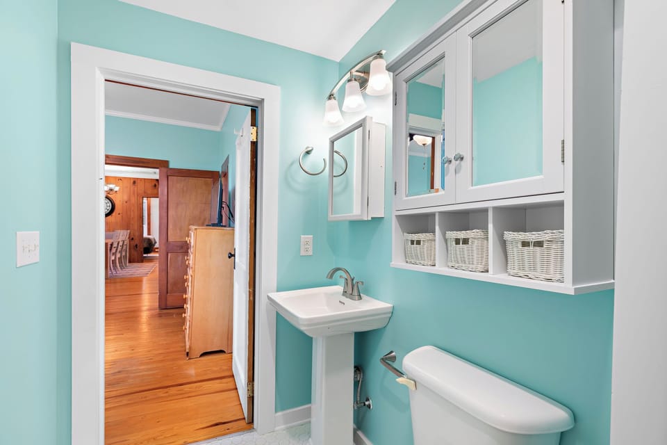 Fresh Oak Island NC bathroom with aqua walls, pedestal sink, mirror, toilet, storage cabinet, and walk-in shower with coastal shell shower curtain and white tile surround.