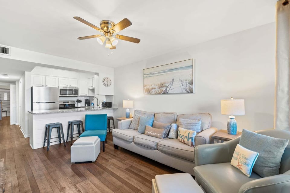 Bright, open-concept living space featuring cozy seating, coastal décor, hardwood floors, and a fully equipped kitchen with bar seating—perfect for relaxing after beach days.