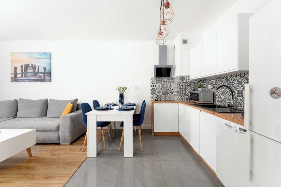 A view toward the kitchen and living area with a white dining table set in the foreground. The patterned backsplash and white cabinetry define the kitchen zone. The gray sofa and wall art complete the living space.