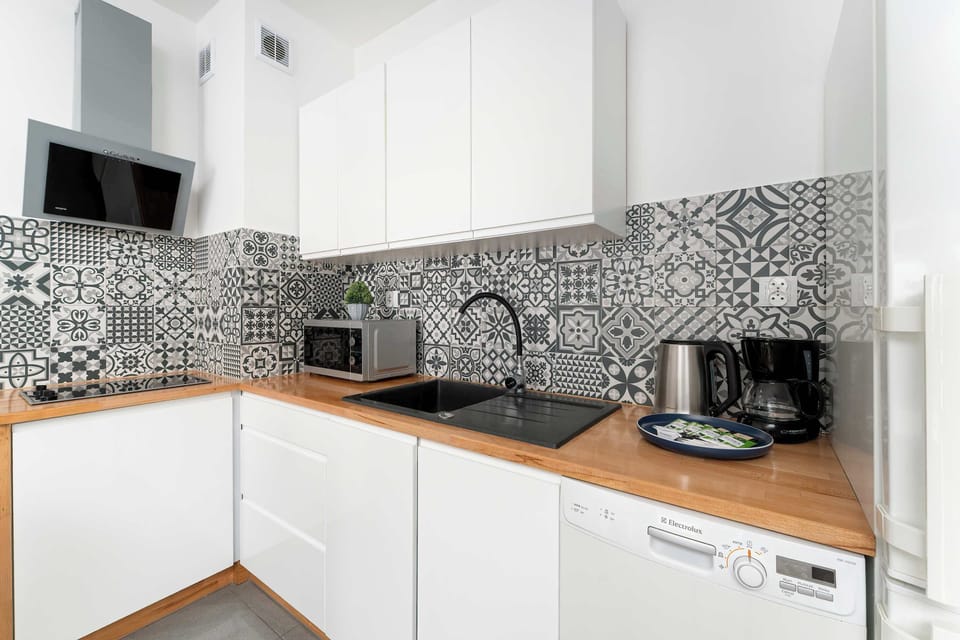 A compact L-shaped kitchenette with white cabinets and a wooden countertop. Patterned black-and-white tiles create a decorative backsplash. Small appliances like a kettle and toaster sit neatly arranged on the counter.