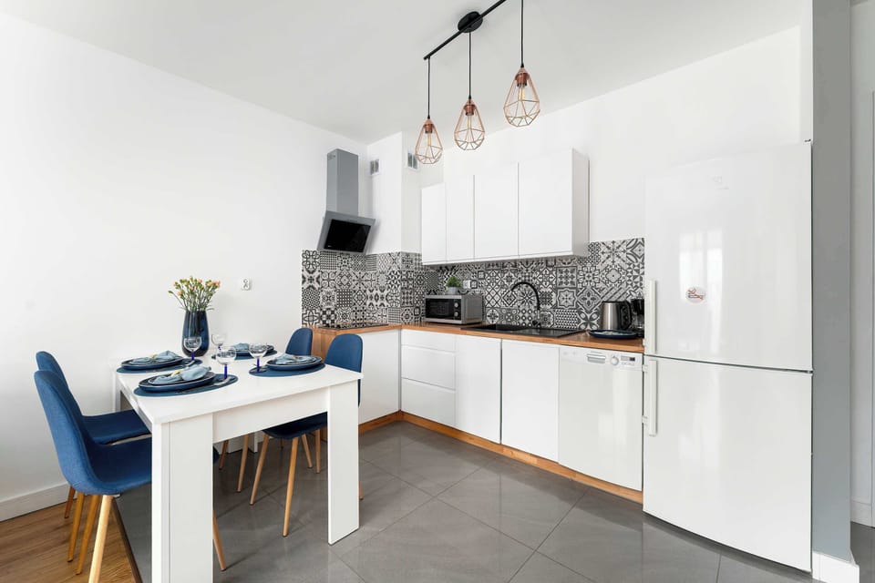 A clean, minimalist kitchen scene with white cabinets and a dining table positioned nearby. Pendant lights hang above the table, adding a decorative touch. The space looks functional and well-organized.