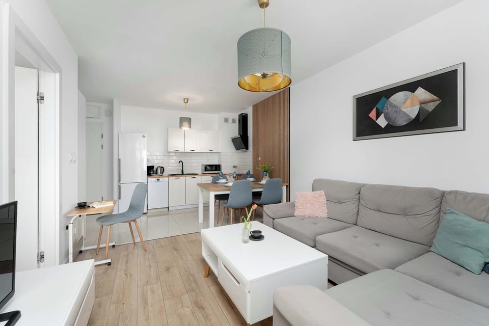 This wide shot captures the flow between the modern kitchen and the inviting living room. The interior design is cohesive, using light wood, white furniture, and blue decorative accents.