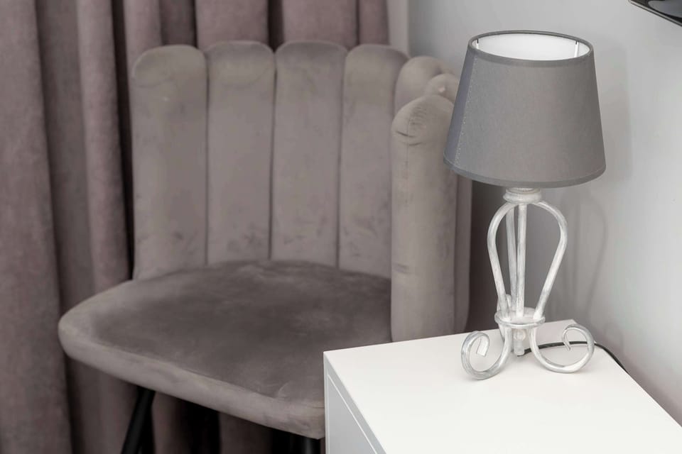 A stylish grey velvet chair sits next to a white bedside table topped with a modern designer lamp. The soft lighting and plush fabrics create a perfect corner for evening reading.