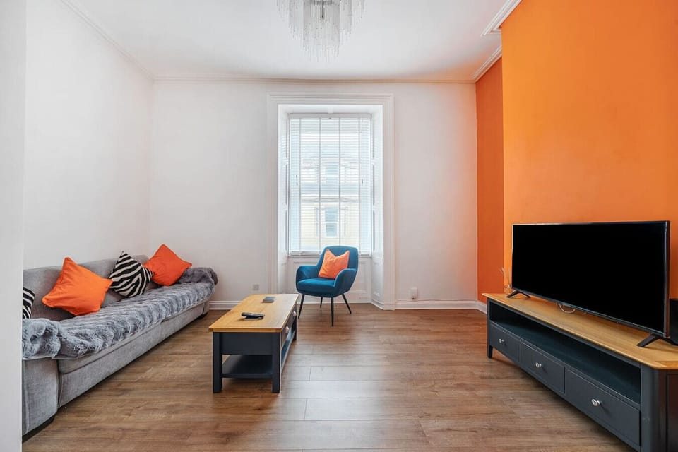 Living room with a grey sofa dressed with orange cushions, a blue armchair, a central coffee table, a large TV on a dark media unit, and a bold orange feature wall opposite the window.