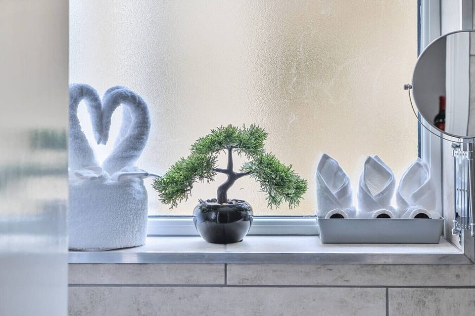 Bathroom windowsill displaying folded towels, a small bonsai plant, and decorative ornaments in front of a frosted window panel.