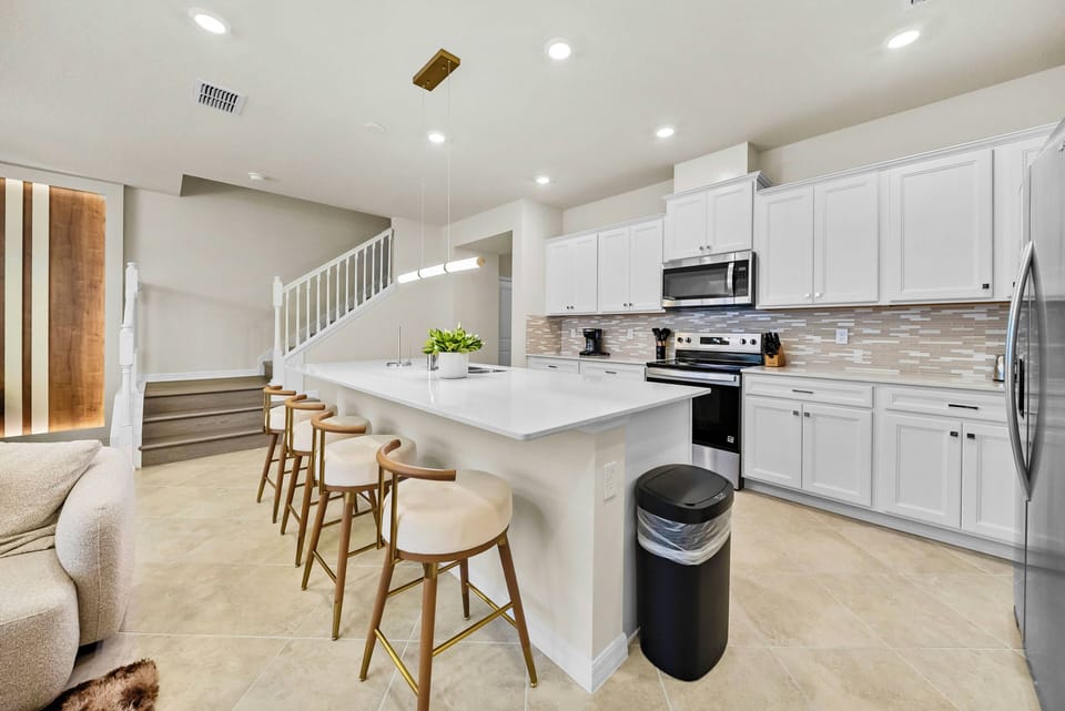 This bright kitchen boasts sleek white cabinetry and a spacious island with stylish bar stools, ideal for casual meals. Modern appliances enhance the warm, inviting atmosphere, making it a perfect spot for cooking and gathering.