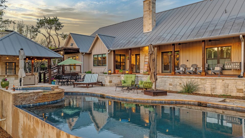 The whole picture. Pool reflecting the evening sky, outdoor kitchen to the left, covered porch to the right, and warm light spilling from every window. Casa Colina at dusk is something else entirely.