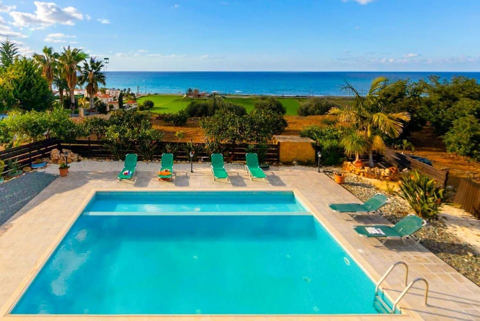 Elevated high-angle view of a private swimming pool with clear turquoise water, green lounge chairs, and a lush garden overlooking the blue ocean coastline under a sunny sky.