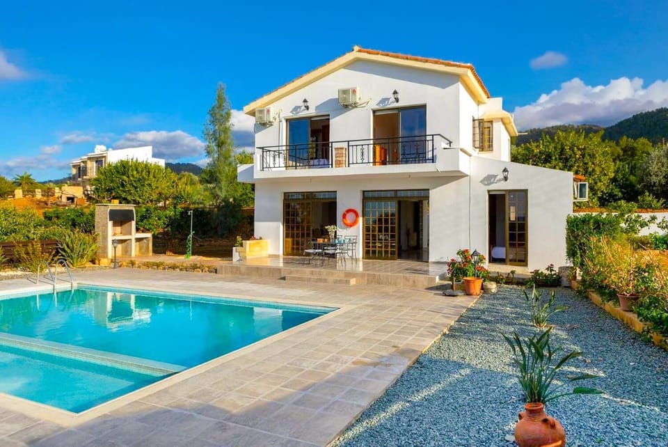 wo-story white holiday villa with an upper balcony, sliding glass patio doors, a private swimming pool, and an outdoor stone barbecue structure under a blue sky.