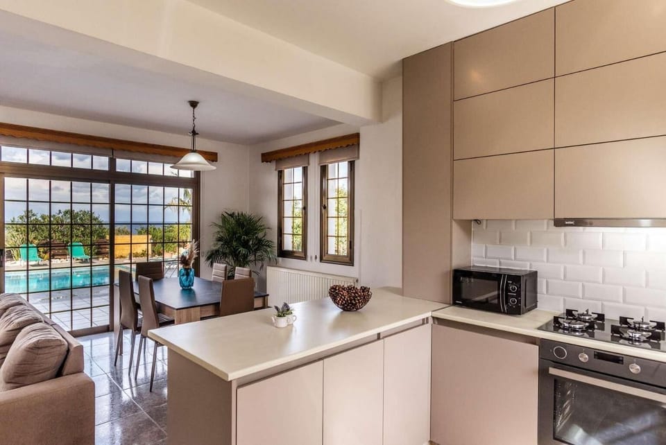Open-plan modern kitchen and dining area featuring minimalist beige cabinets, a breakfast bar, an indoor dining table, a sofa, and sliding glass doors revealing a swimming pool outside.