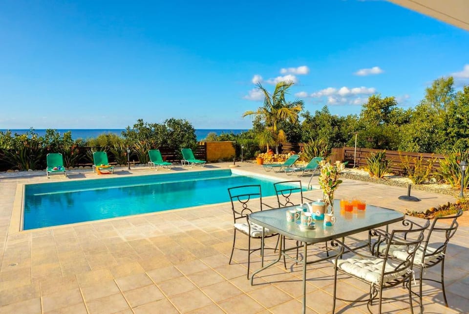 Outdoor glass-top dining table with iron chairs on a patio next to a swimming pool lined with green lounge chairs, offering ocean views in the background.